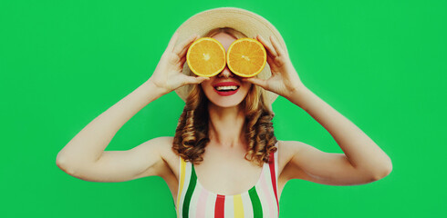 Summer portrait of happy cheerful smiling woman covering her eyes with slices of orange and looking for something wearing a straw hat on green background