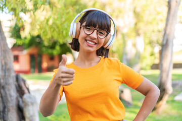 Young brunette woman in the park listening music and with thumb up