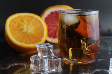 Apple citrus dark juice in a glass next to lies citrus orange grapefruit ice on a black background. Refreshing drinks, summer, soft drinks