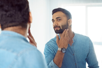 Curious thoughtful indian man touching beard on face looking at mirror