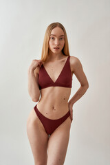 Fototapeta premium Sensual young slim caucasian woman with beautiful perfect body wearing burgundy underwear posing isolated over light gray background