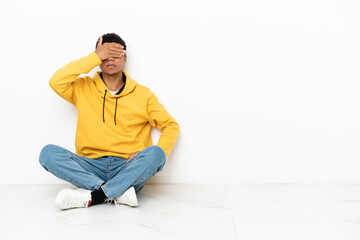 Young African American man sitting on the floor isolated on white background covering eyes by hands. Do not want to see something