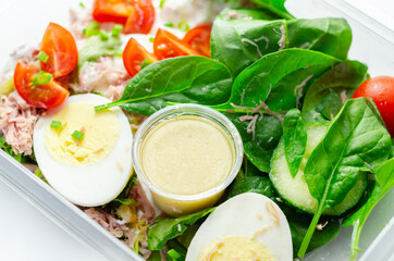 Healthy salad in the plastic food box, potato salad with hard boiled egg, shredded ham, cucumber, tomatoes, lettuce and spinach with a honey and mustard dressing pot