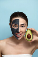 Beautiful young woman with a black mask of clay on face ..Girl model beauty face holding  half an avocado in hand. Cosmetic mask . Skin care . Spa treatment. Cosmetology