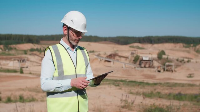 Handsome sand quarry worker engineer, male caucasian foreman in safety wear and hardhat working with digital tablet computer and looking at camera. Bearded industrial worker, mining project specialist