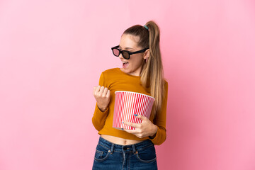 Young Lithuanian woman isolated on pink background with 3d glasses and holding a big bucket of popcorns while looking side