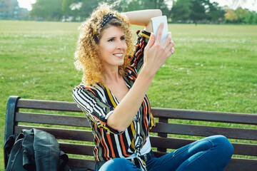 Curly hair woman making selfie photo on a mobile phone. Stylish happy smiling woman in striped multicolor shirt, posing on the background of green lawn. Summer fashion. Capture the moment.