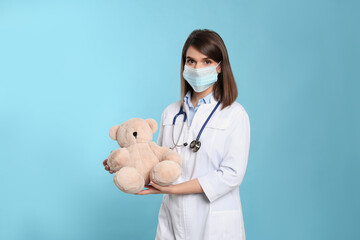 Pediatrician in protective mask with teddy bear on light blue background