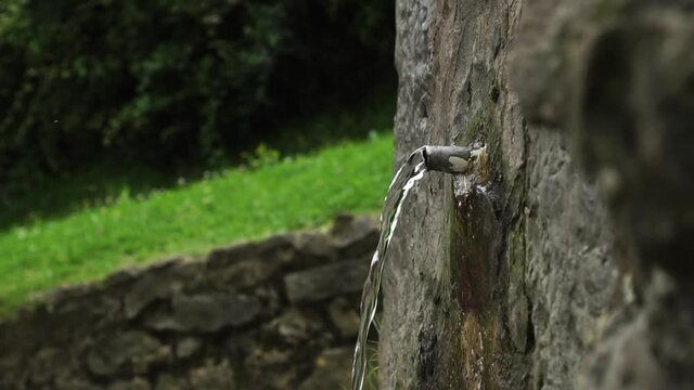 Mineral pure mountain spring with a stone ancient surface. Cold water flows from a metal spout and splashes in slow motion. Vegetation in blur. Close-up