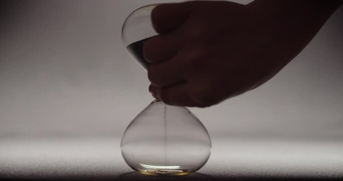 A hand rotating an hourglass with black sand to its cycle beginning. 