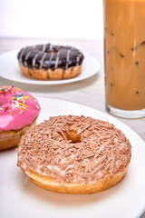 Belgian chocolate chunks donut on a white plate accompanied by an iced latte on a marble surface and a white background with copy space for advertising.