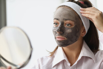 Young woman with black clay mask looking in mirror at home