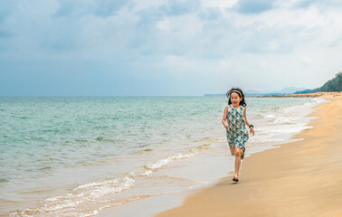 Fototapeta na wymiar Asian little girl running on the beautiful beach, smiling face, black long hair, 7 years old, turquoise color ocean and blue sky. Blank space for text and design.