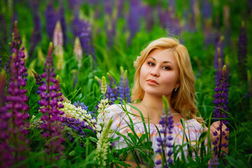 A large portrait of a young plump blonde woman in a blooming field of lupines. A woman is sitting in the grass among tall lilac-pink flowers in a meadow.