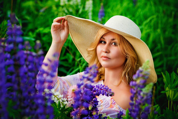 Portrait of a beautiful plump blonde in a large straw hat, a girl with a bouquet of lupines in her hands, posing in a blooming field.