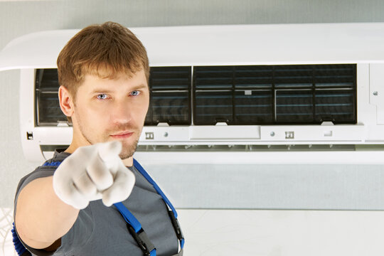 A man in overalls points his finger at the camera against the background of the air conditioner. The concept of reminding you of the need for timely maintenance of the air conditioner