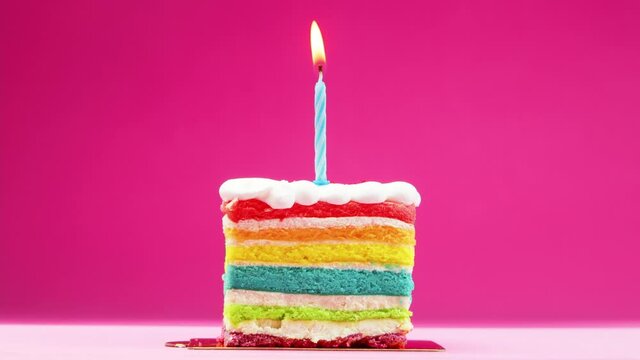 Slow motion, 4K Stock Footage. A slice of birthday cake with a bright blue candle on a pastel pink background. Slow motion, a festive candle is burning on a rainbow piece of cake.