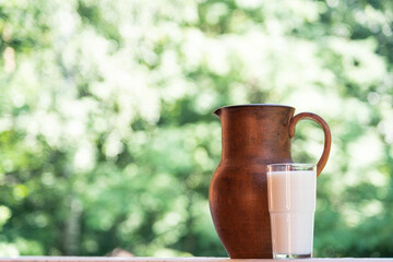 Clay jug and glass of milk on a natural green blurred background. A glass of milk in the summer on...