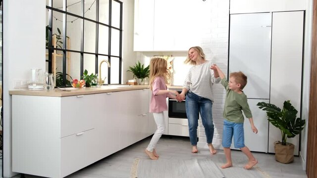 Excited mom or babysiter and two kids boy and girl jumping, dancing laughing in modern scandinavian house kitchen. Happy family mother daughter son having fun enjoy playing singing together at home.