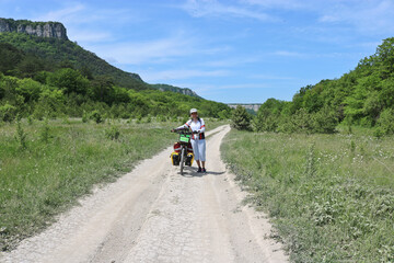 Fototapeta premium A happy cyclist leads her bike along a high plateau among the mountains. Summer landscape, summer bike trip. Mangup-Kale, Crimea.