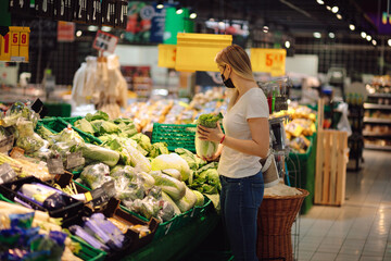 Beautiful young woman in mask chooses vegetables and herbs in supermarket. Proper nutrition. Everyday lifestyle. Natural food. Food products. Personal protective equipment.