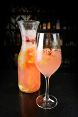 iced pink sangria juice cocktail with rose flower and mixed citrus fruit in jug and glass goblet on black marble bar counter dark night background cold alcohol drink menu