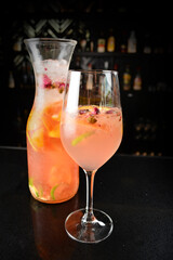 iced pink sangria juice cocktail with rose flower and mixed citrus fruit in jug and glass goblet on black marble bar counter dark night background cold alcohol drink menu
