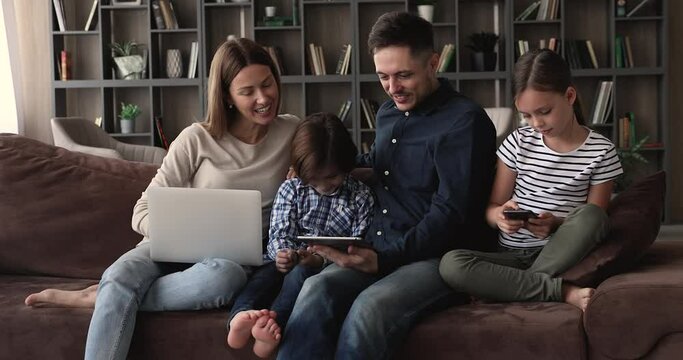 30s couple and little children relax on couch in living room holding laptop smartphone and tablet, using diverse devices. Addicted family, modern gadgets overuse, bad habit of young generation concept