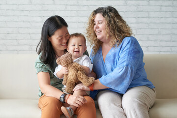 Happy diverse lesbian couple sitting on sofa at home and playing with child