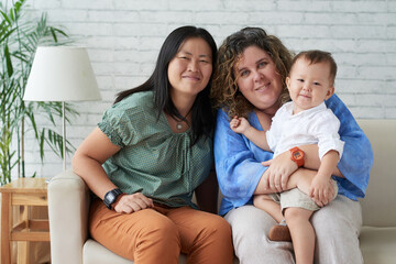 Portrait of smiling lesbian couple sitting on sofa in living room with their little son on laps
