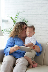 Mother sitting on sofa at home with baby boy, kissing him on cheek and showing animated cartoon on...