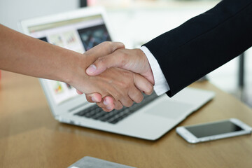 Two businessmen shake hands in an office with computers laptop, smartphone, document on their desks after reaching an agreement to work together to expand.