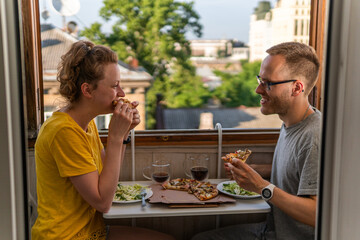 Fototapeta na wymiar A young happy couple is having meal, dinner, enjoying pizza, salad and drinking Kvass at home on the balcony with open windows, amazing view and portable compact table