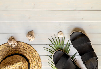 Women's open-toe black sandals from recycled plastic fibers, straw hat, and seashells on the wooden boardwalk. Summer traveling concept. 