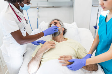 Female doctor talking with patient along coworker in ICU. Man is lying on bed amidst essential workers. Healthcare workers are in protective workwear.