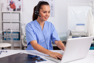 Medical receptionist wearing headset with microphone in private hospital typing on laptop Health care physician sitting at desk using computer in modern clinic looking at monitor.