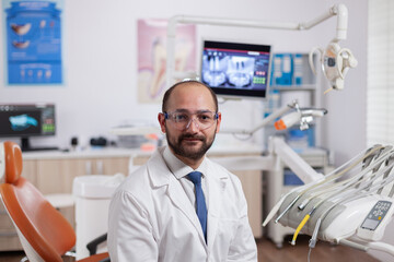 Naklejka na ściany i meble Confident dentist in stomatology cabinet with orange equiptment wearing dental uniform. Medical specialist in oral hygiene wearing lab coat looking at camera in dentistry office.