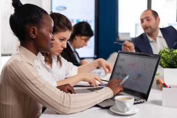 Cheerful african business lady typing on laptop and smiling sitting at desk in busy start up office. African woman using laptop in meeting broadroom, negotiations speaking with clients