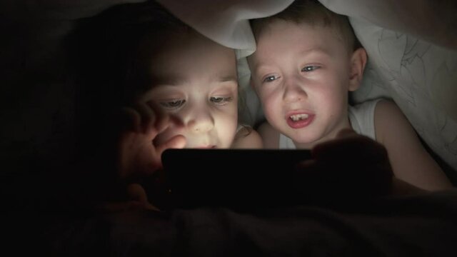 A happy boy and girl lies in bed under a blanket and plays on a phone in a game in the dark. brother and sister face of the child is lit by a bright monitor boy under the covers Computer addict child