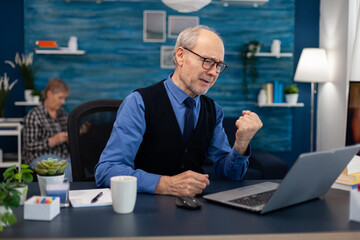 Excited senior man celebrating good news while working on laptop from home office. Elderly man entrepreneur in home workplace using portable computer sitting at desk while wife is reading a book