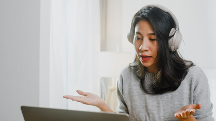 Asia businesswoman wear headphone using laptop talk to colleagues about plan in video call while smart working from home at bedroom. Self-isolation, social distancing, quarantine for corona virus.