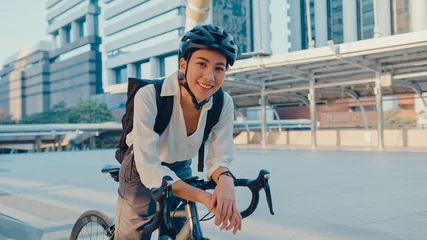 Fotobehang Asian businesswoman go to work at office stand and smiling wear backpack look at camera with bicycle on street around building on a city. Bike commuting, Commute on bike, Business commuter concept. © tirachard