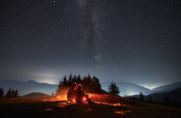 Young man and woman travelers sitting on grassy hill under night sky with stars. Couple hikers looking at majestic night starry sky and hugging. Concept of hiking, night camping and relationships.
