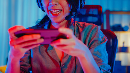 Happy asia girl gamer wear headphone competition play video game online with smartphone colorful neon lights in living room at home. Esport streaming game online, Home quarantine activity concept. © tirachard