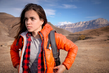 Portrait of a young Caucasian attractive woman standing in a backpack on the background of a rocky mountain peak looks to the side while adjusting her jacket