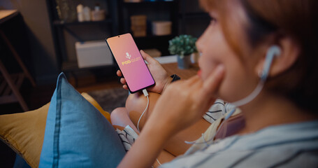 Young attractive Asia lady wear headphones using phone listen digital music podcast online channel sit on sofa in living room at house night. Social distancing, quarantine for coronavirus prevention.