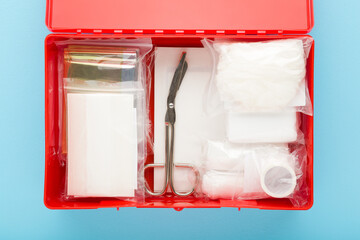 Opened red first aid kit box with different medical accessories in packages on light blue table background. Pastel color. Closeup. Top down view.