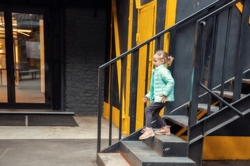 Fototapeta na wymiar Little caucasian blonde girl in fashionable clothes descends the metal steps