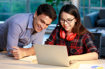 Male and female couple staying in the living room together at evening twilight and working with a notebook computer, discussing with happy faces. New normal, work at home concept.