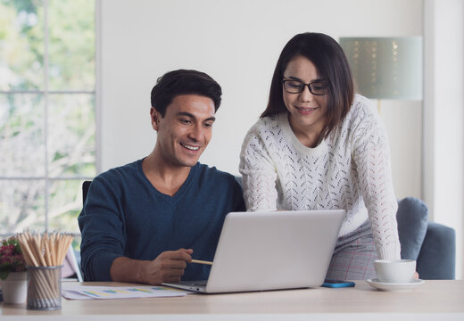 Mixed races couple family, Caucasian man and Asian woman, working together via laptop notebook computer and discussing in living room with happy and intimate. New normal work at home office concept